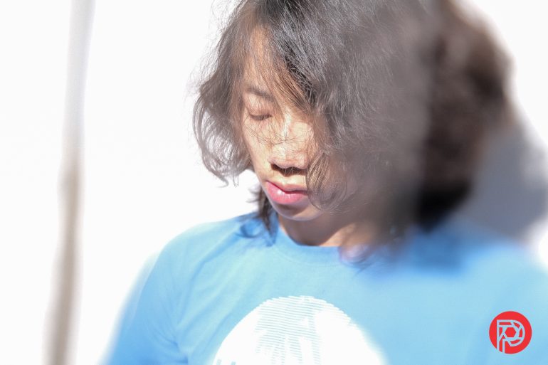 Person with long hair in a blue shirt standing in bright light, eyes closed, with a red logo in the corner.