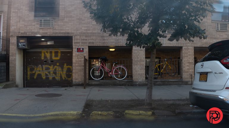 A brick building with “NO PARKING” spray-painted on a garage door, bikes behind a fence, and a parked car.