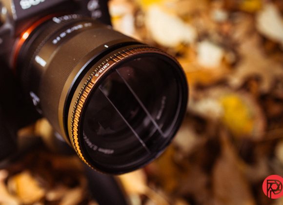 Close-up of a camera lens with a gold filter, surrounded by blurred autumn leaves.