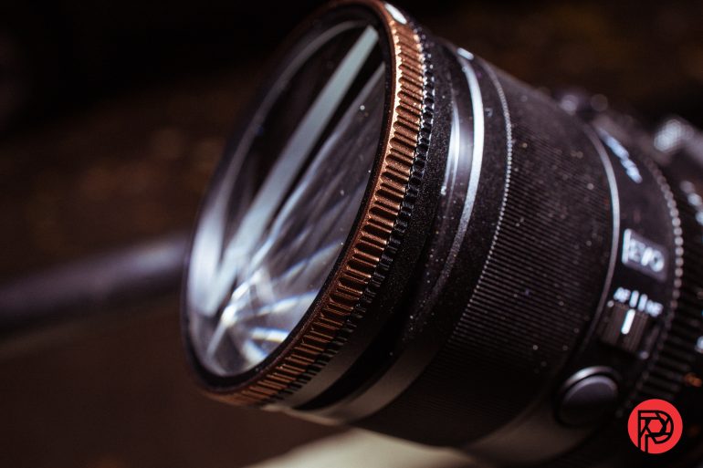 Close-up of a camera lens with water droplets and a red circular logo in the bottom right corner.