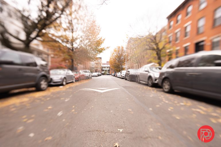 A city street lined with parked cars and autumn trees, viewed from the middle of the road.