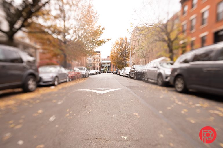 A tree-lined city street with parked cars on both sides and an arrow painted on the road.