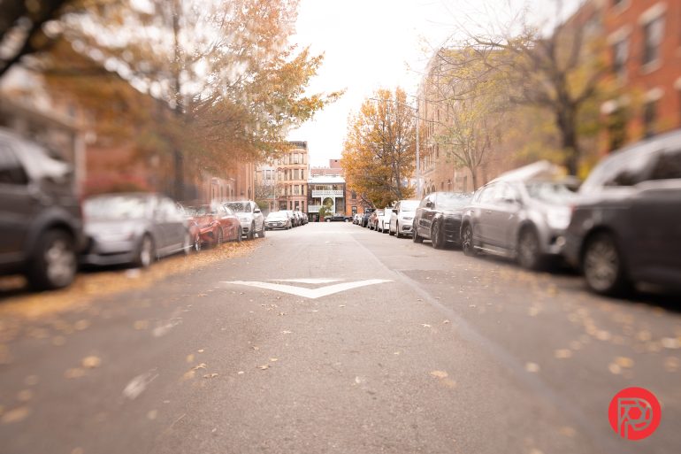 A city street lined with parked cars and autumn trees, viewed from ground level with a white arrow on the asphalt.