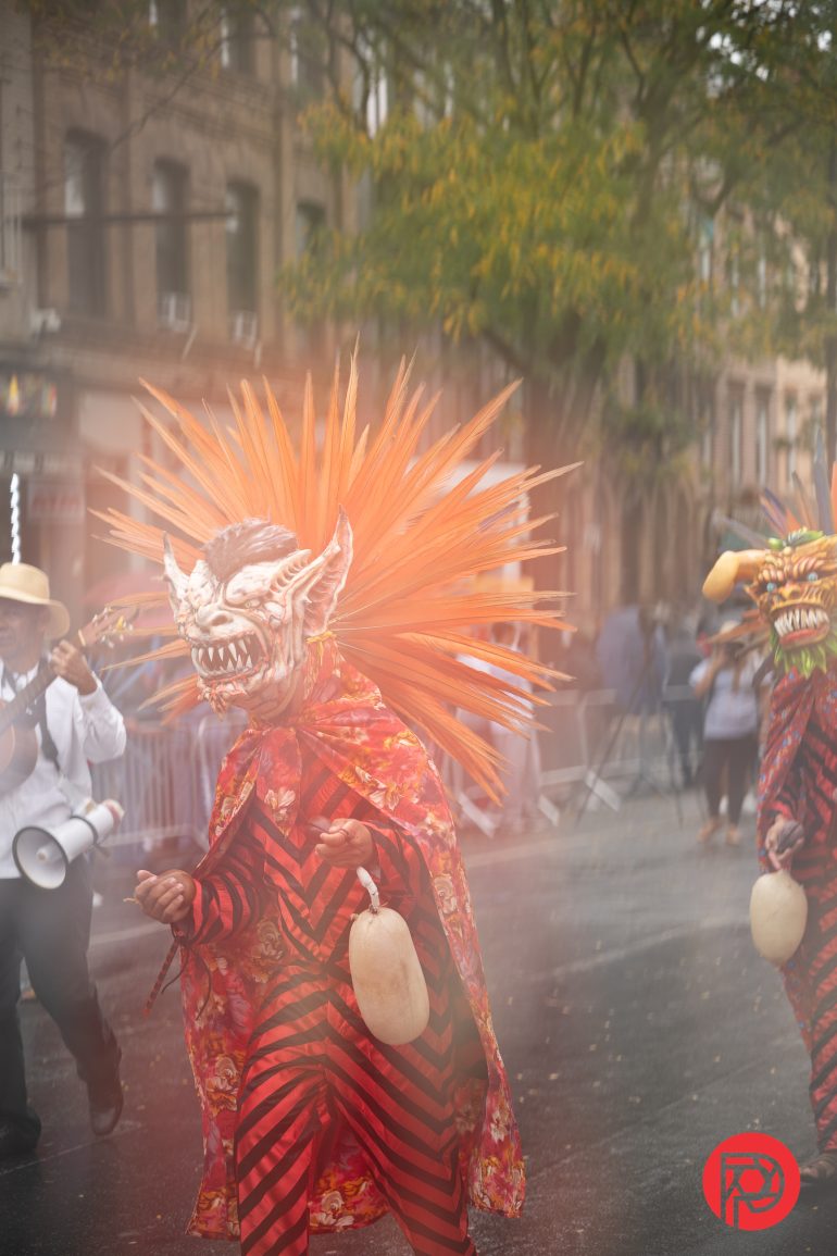 Person in a vibrant costume and spiked mask walks in a street parade, holding a rattle, with blurred onlookers.