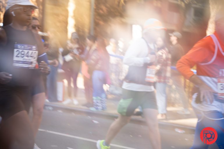 Runners in a race move along a sunlit street, with blurred spectators and sunlight in the background.