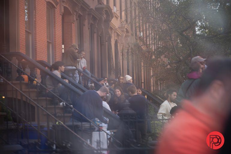 People gather and socialize on brownstone stoops along a city street on a hazy day.