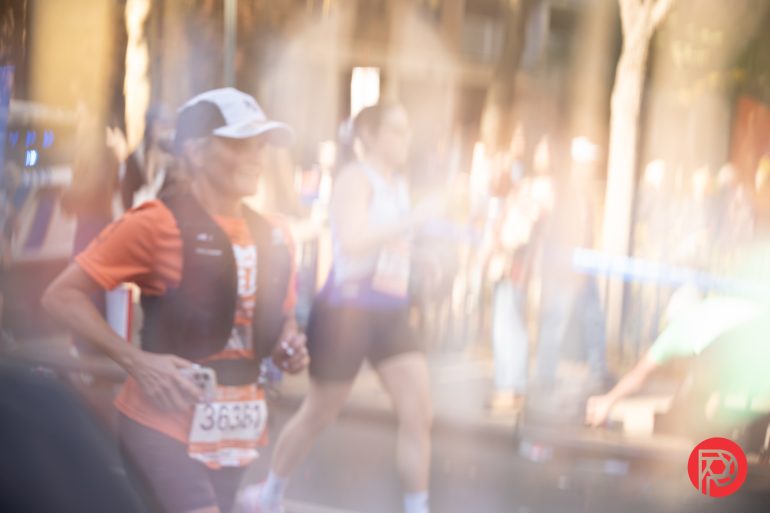A blurred image of marathon runners, with one runner in focus wearing a cap and orange shirt.