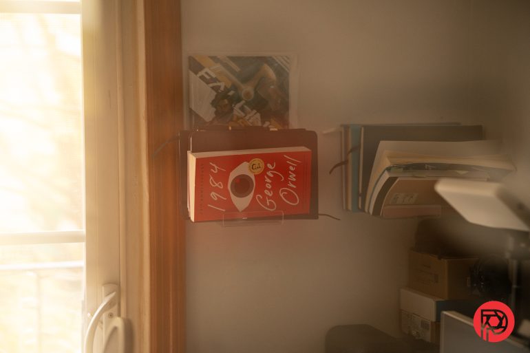 A copy of George Orwells 1984 is displayed on a shelf next to other books in a softly lit room.