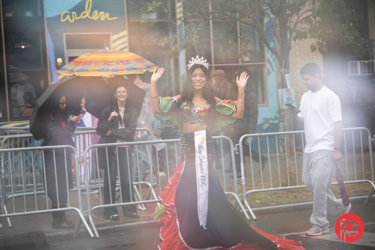 A smiling woman in a crown and sash waves in a parade, with people and umbrellas in the background.