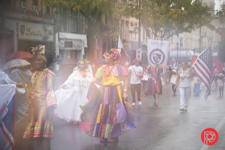 People in colorful traditional clothing walk in a parade on a rainy street, some holding flags and banners.