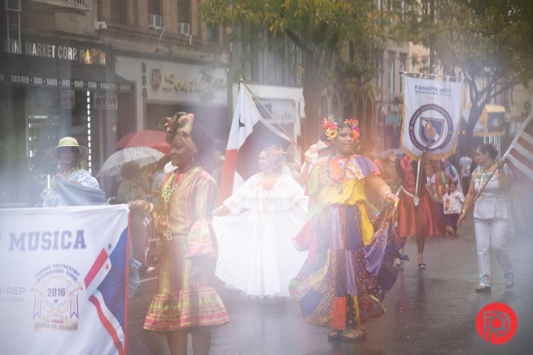 People in colorful traditional clothing march in a parade on a rainy city street, carrying banners and flags.