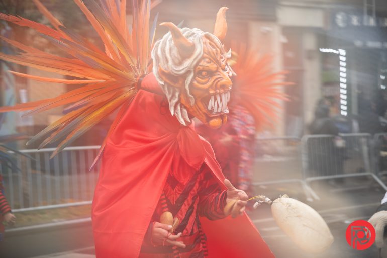 Person in an orange demon mask and costume with wings, holding a drum, at an outdoor parade or festival.