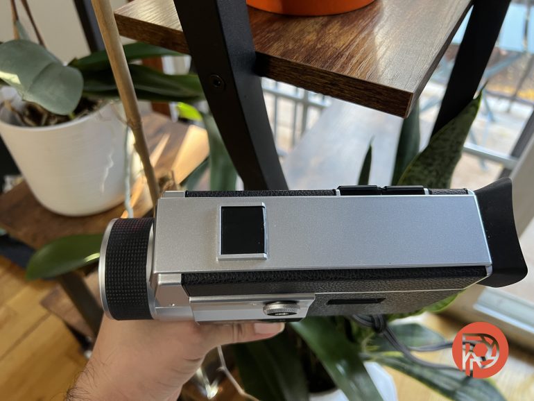 A hand holds a vintage camera near indoor plants by a window and wooden shelf.