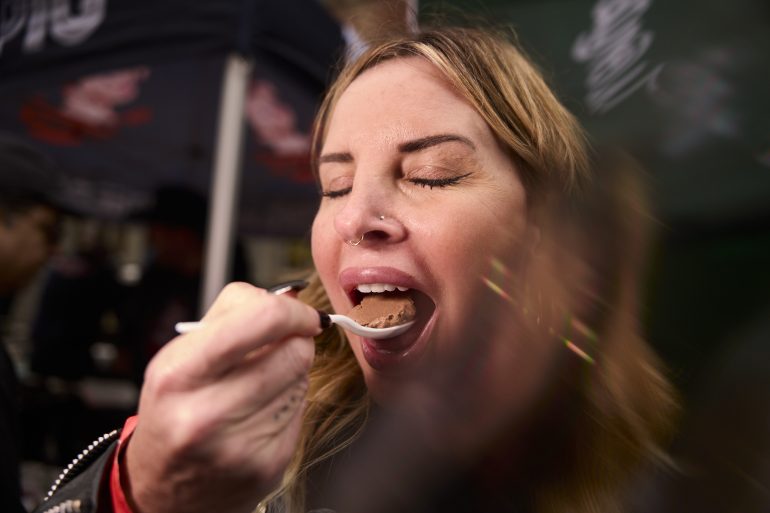 Woman with closed eyes eating food from a spoon, appearing to enjoy the taste, with blurred background.