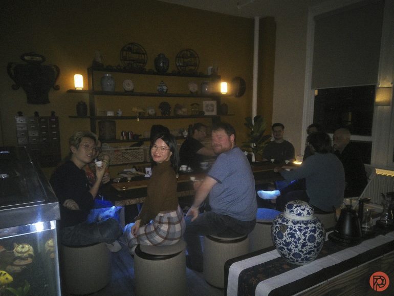 A group of people sit around a cozy, dimly lit tea room, smiling and enjoying tea together.