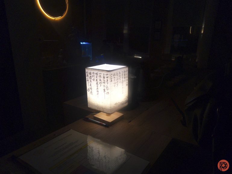 A lit paper lantern with Japanese writing on a wooden table in a dimly lit room.