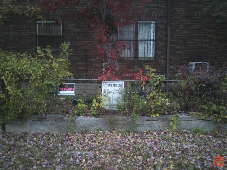 A brick house with autumn leaves, a No Trespassing sign, and another sign by a tree in the yard.
