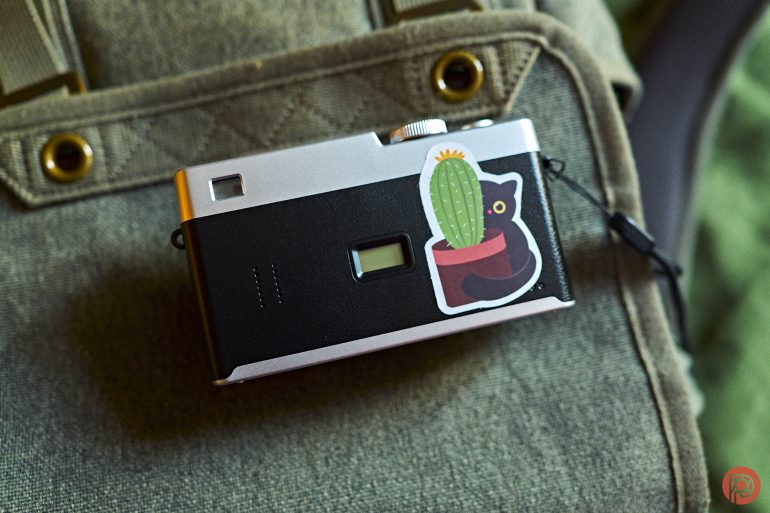A camera with a cat and cactus sticker rests on a khaki bag.