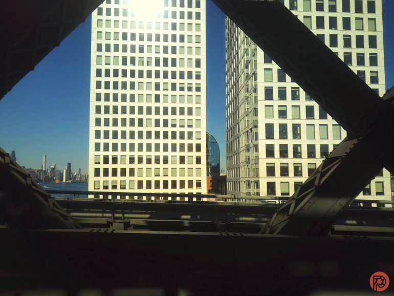 View of two tall white office buildings framed by bridge beams, with a city skyline in the background.
