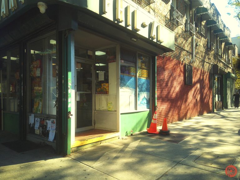 A small corner store entrance with two orange traffic cones on the sunlit sidewalk.