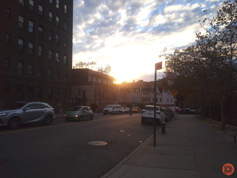 Cars parked along a city street at sunset, with buildings and trees on both sides under a cloudy sky.