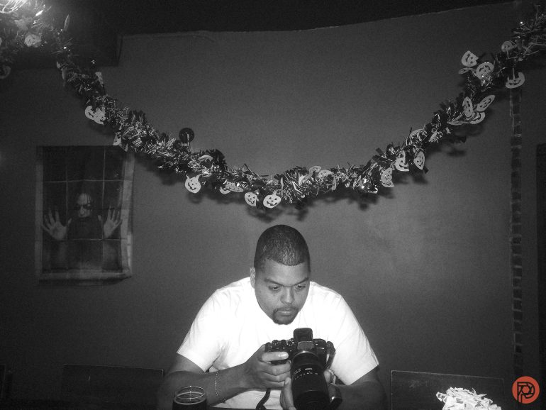 A man looks at a camera in a dim room decorated with Halloween garland and a spooky poster on the wall.