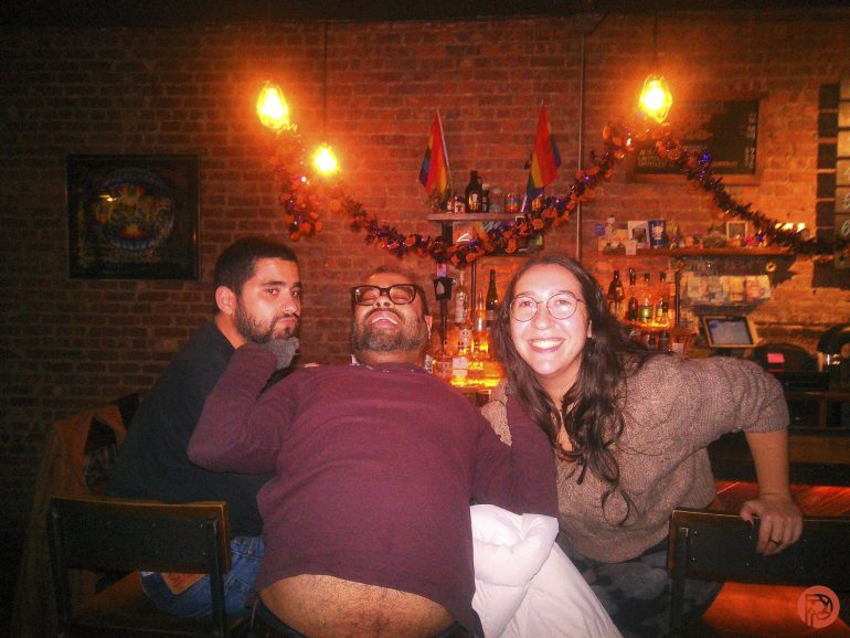 Three friends smiling and posing together at a cozy, warmly lit bar with festive string lights.
