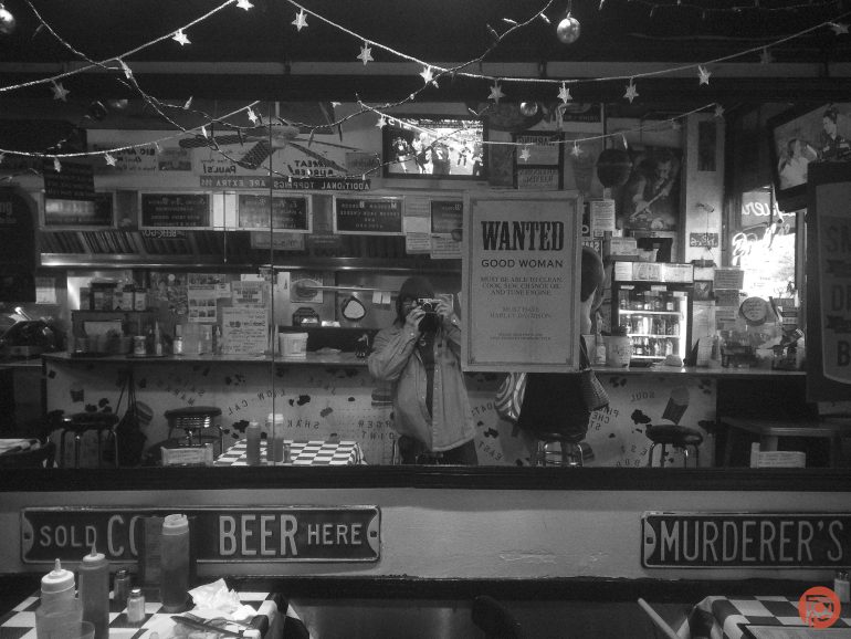 A person takes a photo in a diner with eclectic signs, string lights, and a WANTED poster on the mirror.