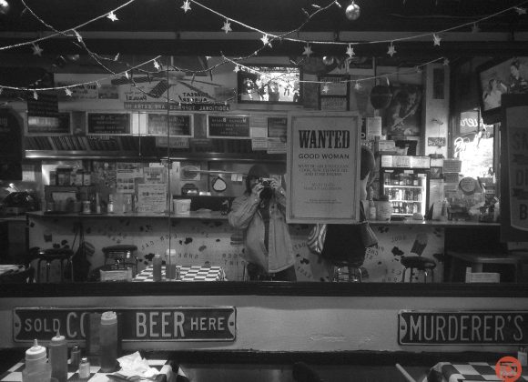 Person taking a photo in a diner with vintage signs, string lights, and a WANTED poster reflected in a mirror.