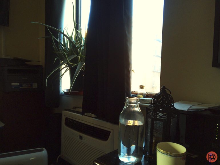 A glass jar of water, a cup, and a lantern sit on a table by a window with a plant.