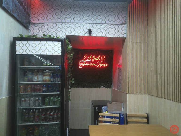 Small eatery with a drinks fridge, tables, and a neon sign reading Eat Fresh!!! Shawarma House on the wall.