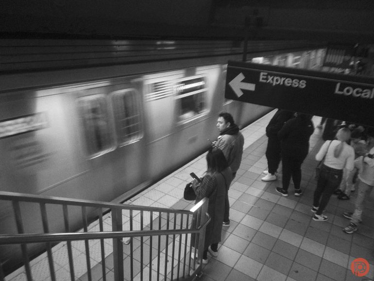 Black and white photo of people waiting on a subway platform as a train speeds by.