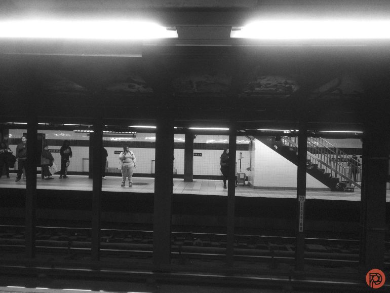 Black and white photo of people waiting on a subway platform with stairs visible on the right.