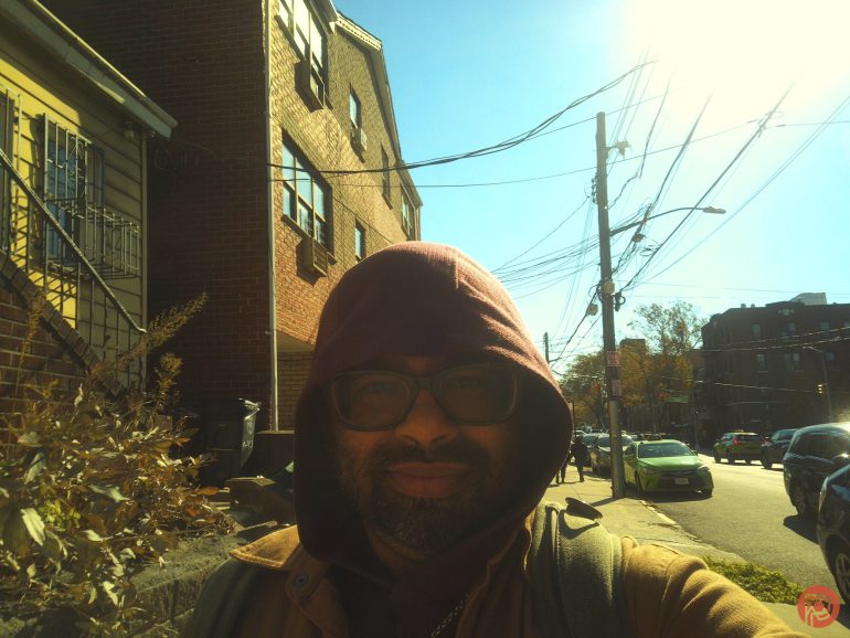 A person in a hooded jacket and glasses smiles on a sunny street with houses and parked cars.