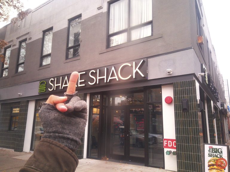 A person in gloves holds up their middle finger in front of a Shake Shack restaurant.