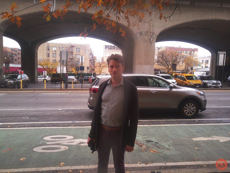 A man stands on a city sidewalk under an overpass, with cars, buildings, and autumn trees in the background.
