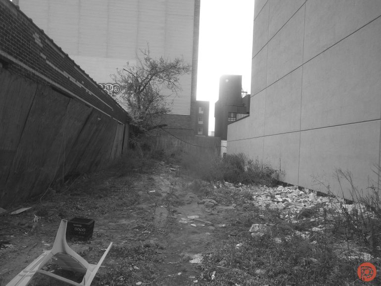 Vacant lot between buildings with overgrown weeds, scattered trash, and a broken white chair in the foreground.