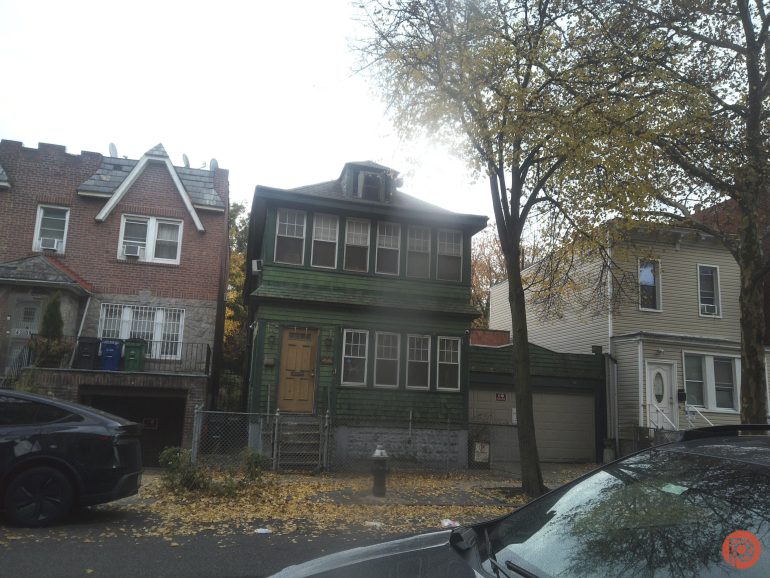 A green two-story house with boarded windows, surrounded by trees and parked cars on a city street.