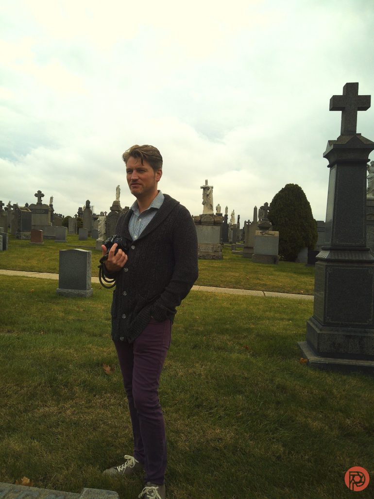 A man stands in a cemetery holding a camera, surrounded by gravestones under a cloudy sky.