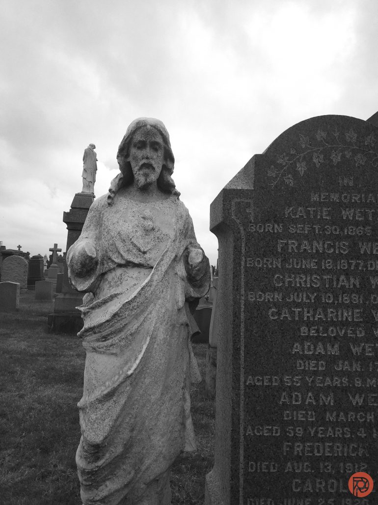 A weathered statue of Jesus stands beside an engraved gravestone in a cloudy cemetery.