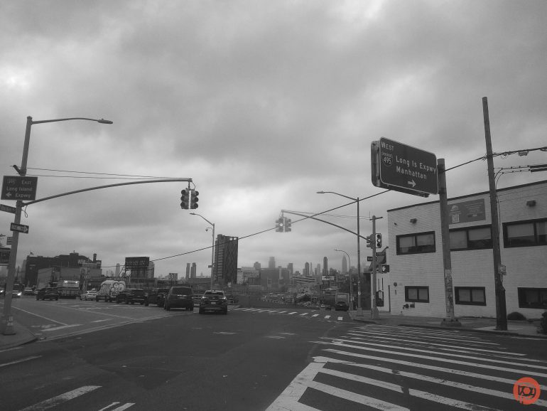 A city street under cloudy skies, with traffic lights, street signs, and Manhattan skyline in the distance.