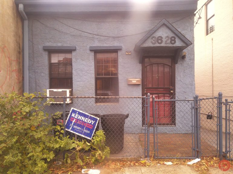 A small blue house with a fenced yard and a tilted Kennedy for Judge sign by the gate.