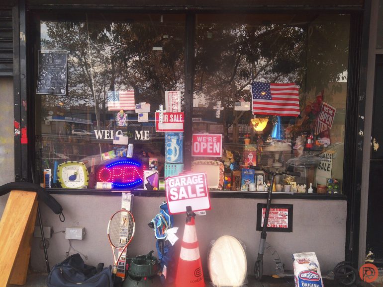 A storefront window with garage sale signs, assorted items, and an American flag displayed.