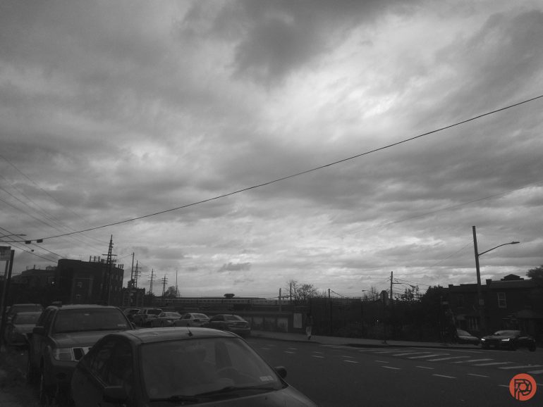A cloudy sky over a city street lined with parked cars and buildings in black and white.