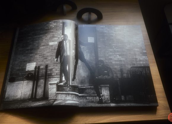 Open photobook on a wooden table showing two men near a brick building, one seated and one standing outside.