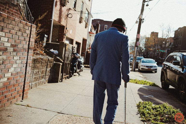 An elderly person in a blue suit walks on a city sidewalk with a cane on a sunny day.