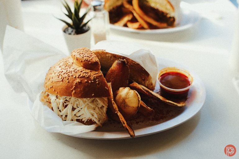 A sandwich with shredded cabbage, chips, a fried item, and sauce on a white plate next to a small plant.