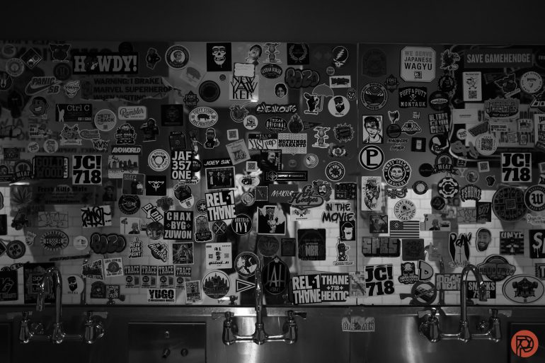 A wall covered in various stickers above a row of metal sinks in a dimly lit room.