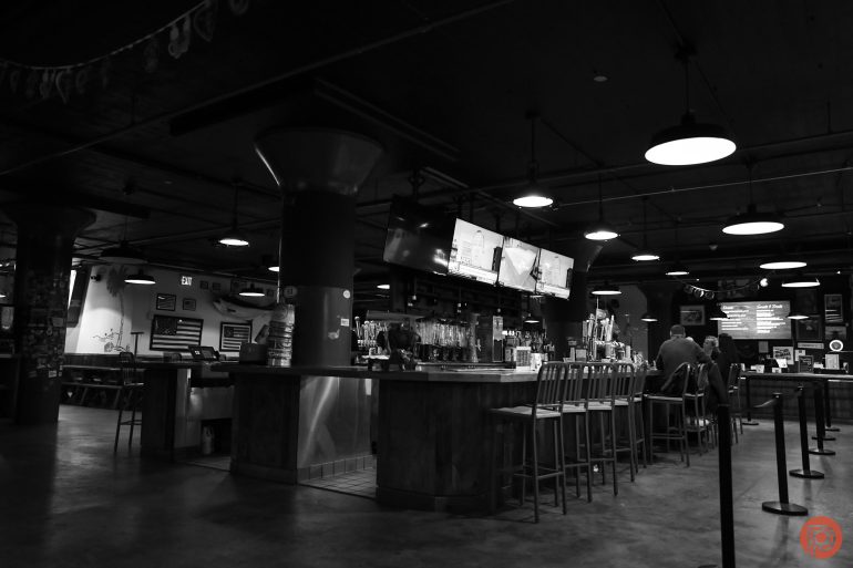 Black and white photo of a bar with empty stools, TVs overhead, and a few people sitting at the counter.