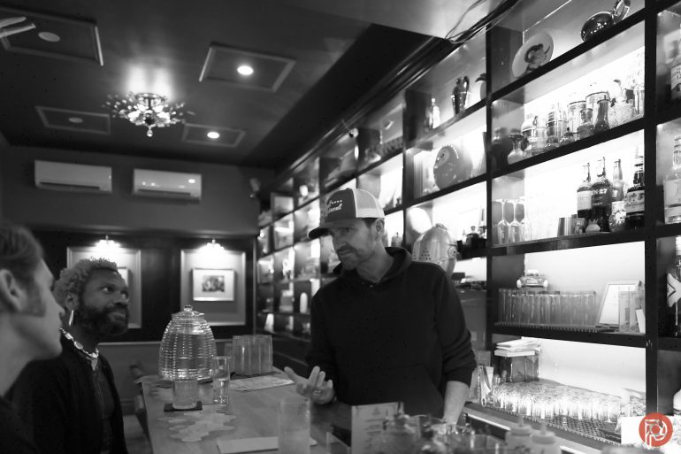 A bartender talks to two customers at a bar with shelves of glasses and bottles behind him. Black and white photo.